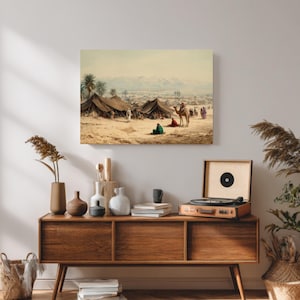 Puede incluir: Una impresión antigua representa una escena desértica con tiendas de campaña, palmeras y figuras, incluida una persona montada en un camello. La obra de arte está enmarcada y montada en una pared blanca sobre un mueble de madera.