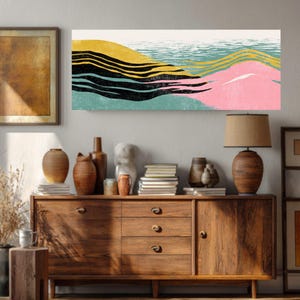 May include: Abstract landscape art print featuring a colorful design. The artwork showcases a layered composition with yellow, black, pink, and teal hues, evoking a sense of rolling hills or waves. The print is displayed above a wooden cabinet with various decorative items.