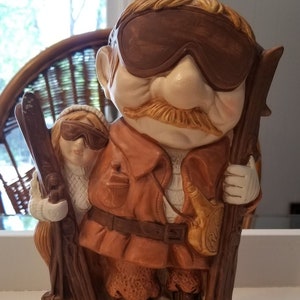 May include: A ceramic figurine of a man and woman wearing ski goggles and holding skis. The man is wearing a brown jacket and the woman is wearing a white sweater. The figurine is standing on a base that says "SNOW BIRDS".