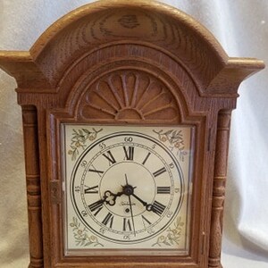 May include: A wooden mantel clock with a white face and black Roman numeral hour markers. The clock has a decorative floral design around the edge of the face.
