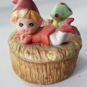 May include: A ceramic trinket box featuring a small elf figurine lying down with a red hat and a red and white outfit. The elf is next to a green mushroom with red dots. The box is decorated with a brown, textured surface that resembles tree bark.