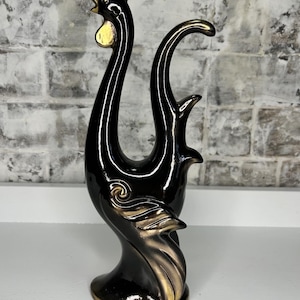 May include: A black and gold ceramic rooster figurine. The rooster has a sleek, elongated design with a glossy black finish and gold accents on the comb, wattle, and tail feathers. The figurine is standing on a small, round base.