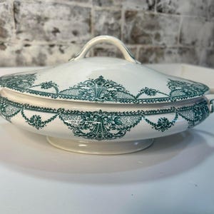 May include: White ceramic serving dish with a lid, featuring a green floral pattern around the rim and on the lid. The dish has two handles and a handle on the lid. The dish is on a white surface.