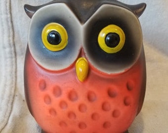 May include: A red ceramic owl piggy bank with black and yellow details. The owl has large, round eyes with yellow pupils and a small, yellow beak.