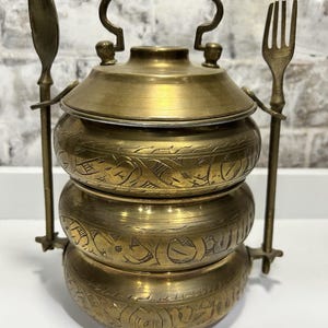 May include: A three-tiered brass tiffin carrier with intricate engraved designs. The carrier has a lid and comes with a spoon and fork.