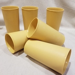May include: Six yellow plastic tumblers, stacked and arranged on a white surface. The tumblers are a pale yellow color and have a smooth, rounded shape.