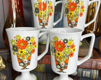 May include: Set of four white ceramic mugs with a floral design. The mugs feature a bouquet of orange, yellow, and green flowers in a green pot. The mugs have a pedestal base and a green rim.