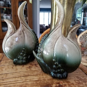 May include: Two ceramic vases shaped like stylized green and white flowers with textured details. The vases are sitting on a wooden surface.