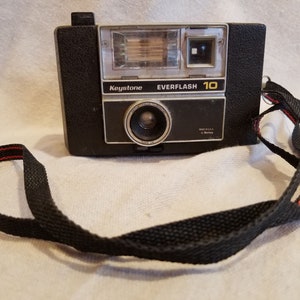 May include: A black Keystone Everflash 10 camera with a built-in flash and a black strap.
