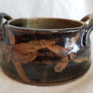 May include: A handmade ceramic bowl with two loop handles. The bowl is dark brown with orange and tan abstract patterns. The interior is a lighter brown. The bowl is likely used for serving or display.