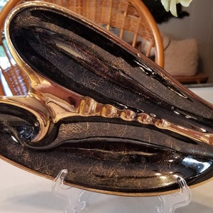 May include: A black and gold ceramic ashtray shaped like a lobster. The ashtray has a gold-colored interior and a black exterior with gold accents. The ashtray is sitting on a clear plastic stand.
