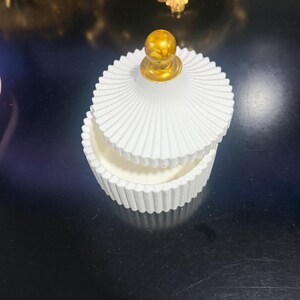 以下が含まれることがあります： ベースと蓋の両方にリブ状の溝があるテクスチャーが施された、白い円形の蓋付き容器。蓋の中央には、滑らかな金色の小さな球状のつまみが付いています。容器は上からのアングルで撮影されています。
