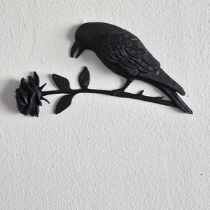 May include: A black decorative wall hanging featuring a raven perched on a rose branch. The raven is detailed with textured feathers and a curved beak. The rose is in full bloom, with detailed petals and leaves.