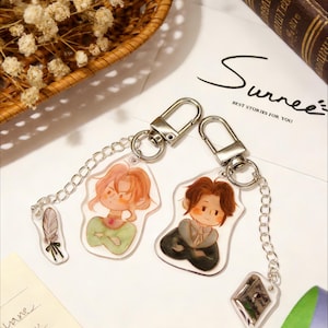 May include: Two keychains with cartoon illustrations of characters. One keychain features a feather charm. The keychains are attached to silver-colored clasps and chains. The background includes a book, dried flowers, and a handwritten note.