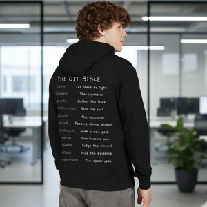 Op de afbeelding: Zwarte hoodie met witte tekst op de rug. De tekst luidt "THE GIT BIBLE" bovenaan, gevolgd door een lijst met git-opdrachten en hun bijbehorende zinnen. De persoon draagt de hoodie.