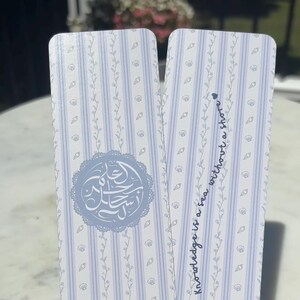 May include: Two rectangular bookmarks featuring a light blue and white vertical striped pattern with small seashell and vine illustrations. One bookmark displays Arabic calligraphy in a circular frame, while the other reads Knowledge is a sea without a shore.