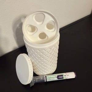 May include: A white, textured container with a lid featuring multiple circular openings. The lid is off, revealing the interior. A pen-like medical device labeled "zepbound" lies next to the container.
