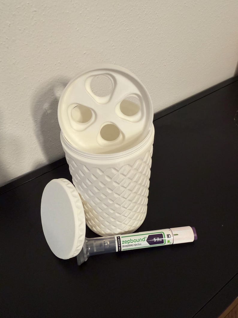May include: A white, textured container with a lid featuring multiple circular openings. The lid is off, revealing the interior. A pen-like medical device labeled "zepbound" lies next to the container.