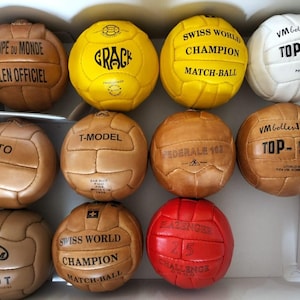 Puede incluir: Una colección de balones de fútbol vintage en varios colores, incluyendo marrón, amarillo, blanco y rojo. Cada balón presenta textos y diseños únicos, como "COUPE DU MONDE" y "SWISS WORLD CHAMPION". Los balones están dispuestos en una caja.
