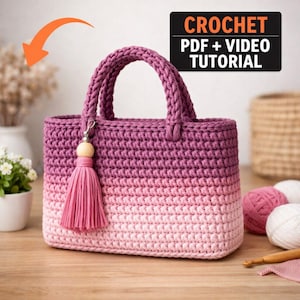 May include: A handmade crochet tote bag in a gradient of pink hues, with a matching tassel and wooden bead charm. The bag features a sturdy handle and is displayed on a wooden surface. The image includes text that reads "CROCHET PDF + VIDEO TUTORIAL".