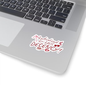 May include: A white sticker with red text that reads "Radicalized BY BASIC DECENCY!" The design includes a bow, hearts, and an arrow. The sticker is placed on a silver laptop.