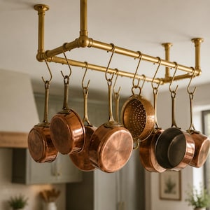 May include: A rectangular brass ceiling-mounted pot rack with multiple S-shaped hooks. Hanging from the hooks are various copper-toned cookware pieces, including several saucepans, a small colander, and a frying pan, all featuring brass handles.