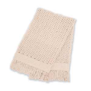 May include: Cream-colored knitted throw blanket with a chevron pattern. The blanket has a fringed edge and is folded. The soft, neutral color and textured design make it a cozy home decor item.