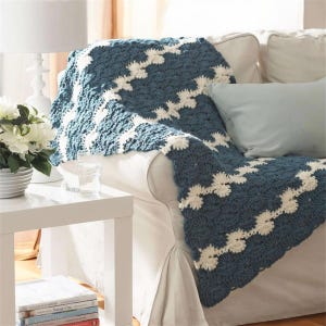 May include: A blue and white crochet blanket draped over a white sofa. The blanket features a repeating zigzag pattern in a dark blue and cream color. A light blue pillow rests on the sofa. A white side table with a lamp and flowers is in the background.