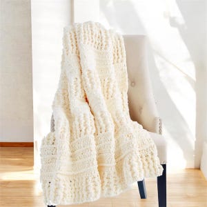 Może przedstawiać: Kremowy, gruby dzianinowy koc narzucony na beżowe krzesło z tapicerką. Koc ma wzór o fakturze z poziomymi i pionowymi żebrami. Krzesło ma pikowane oparcie i ciemnoniebieskie nogi, tworząc przytulną, zachęcającą estetykę.