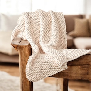 May include: A cream-coloured, chunky knit throw blanket draped over a rustic wooden bench. The blanket's texture is visible, with a dense, raised stitch pattern. The neutral colour and cosy appearance suggest comfort and warmth.