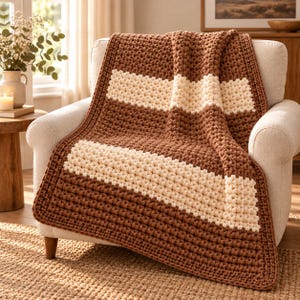 May include: A chunky crochet throw blanket featuring a textured bobble stitch pattern. The blanket is designed with alternating horizontal stripes in solid brown and cream colors, finished with a thick, uniform border around the entire edge.