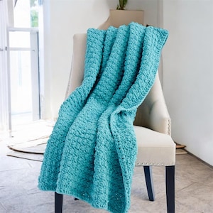 Puede incluir: Una manta turquesa texturizada sobre una silla color crema. La manta tiene un aspecto grueso y afelpado, con costuras visibles y una textura suave y acogedora. La silla tiene respaldo con botones y patas oscuras, sobre un fondo de luz natural.