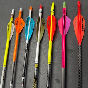 Könnte beinhalten: Eine Sammlung von sieben Pfeilen mit verschiedenfarbigen Befiederungen, darunter Gelb, Orange, Blau, Grün, Pink und Rot. Die Pfeile haben schwarze Schäfte mit den Texten "BADASS", "AAE HYBRID", "ALRAZA" und "ADE MAX".