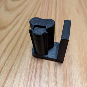 May include: A black 3D-printed object, likely a holder or stand, featuring a cylindrical component with a curved top and a rectangular base. The design includes a vertical support and a slot, suggesting a specific function.