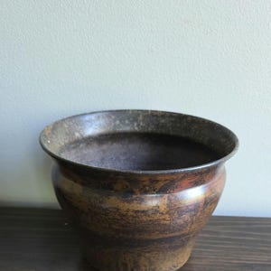May include: A rustic, antique-style planter pot with a wide, slightly flared rim. The pot has a dark, mottled finish with shades of brown and bronze, giving it a weathered appearance. The interior is dark, and the pot sits on a dark wooden surface.