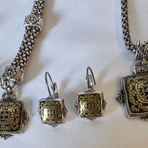 May include: A silver-toned jewelry set featuring a necklace, bracelet, and earrings. Each piece has a square pendant with a gold-colored filigree design on a black background. The bracelet and necklace have a chain-like design.