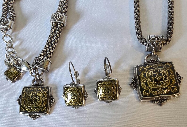 May include: A silver-toned jewelry set featuring a necklace, bracelet, and earrings. Each piece has a square pendant with a gold-colored filigree design on a black background. The bracelet and necklace have a chain-like design.