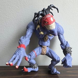 May include: A blue action figure with a red face and white teeth. The figure has black appendages on its head and red accents on its limbs. It is posed on a wooden surface.