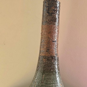 May include: A tall, slender ceramic vase with a textured, uneven surface. The base is a muted, speckled sage green, transitioning into a band of earthy terracotta brown, and finishing with a dark, glossy brown neck and rim. The form is organic.
