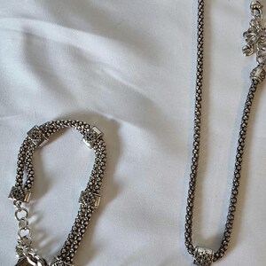 May include: A silver-toned jewelry set featuring a necklace, bracelet, and earrings. The necklace and bracelet have a chain-like design with square accents. The earrings and pendant have a gold-colored filigree design.