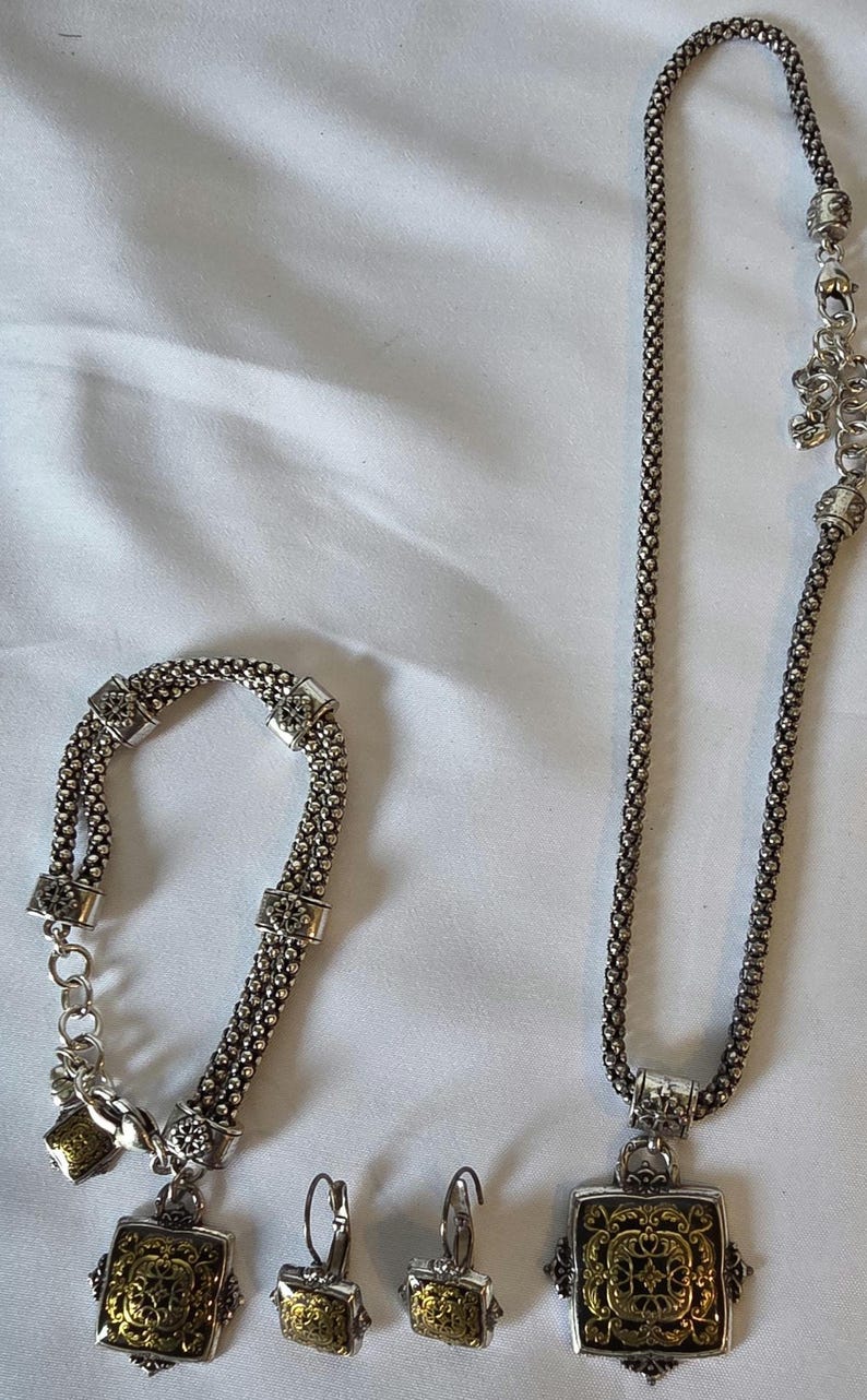 May include: A silver-toned jewelry set featuring a necklace, bracelet, and earrings. The necklace and bracelet have a chain-like design with square accents. The earrings and pendant have a gold-colored filigree design.