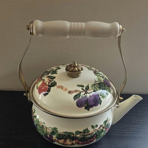 May include: A cream-colored enamel tea kettle featuring a colorful fruit pattern of grapes, plums, and apples. The kettle has a brass-toned metal handle with a white ceramic grip, a matching brass-toned lid knob, and a short, angled spout.
