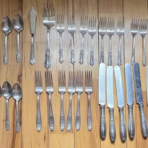 May include: An assortment of vintage silverware, including spoons, forks, and knives, arranged on a wooden surface. The silverware has a silver finish and ornate handle detailing. The collection includes various sizes and styles of utensils.