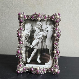 May include: A rectangular picture frame decorated with purple and white beads and small crystals. The frame holds a black and white photograph of three children, with the text "Yes, it's Prada and yes, it's real!" printed at the bottom.