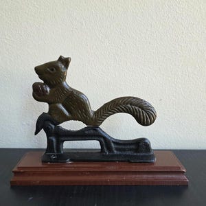 May include: A cast iron nutcracker shaped like a squirrel holding a nut, featuring a bronze-toned squirrel figure mounted on a black metal base, which is attached to a rectangular, dark brown wooden platform for stability and display.