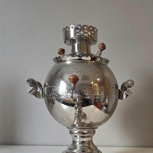 May include: A silver-colored samovar with a round body and ornate details. It features a spigot, wooden handles, and a decorative top. The samovar sits on a pedestal base with three feet. The word "Водолей" is engraved on the body.