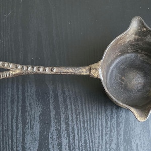 May include: Antique cast iron ladle with a long handle and a round bowl with three spouts. The handle has the text "M. ROBERTS & SON, RACINE AUG 15 1871". The ladle is dark gray with signs of wear.