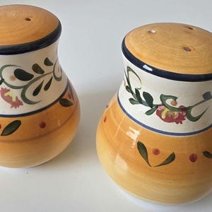 May include: A pair of ceramic salt and pepper shakers. The shakers have a yellow base and a cream-colored middle section with floral and leaf designs. The tops are yellow with a dark blue rim.