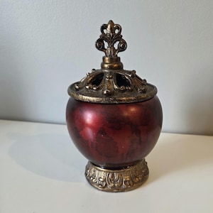 May include: A decorative jar with a red and gold finish. The jar has a round body, a decorative lid, and a detailed base. The lid is adorned with an ornate finial. The jar is likely made of ceramic or a similar material.