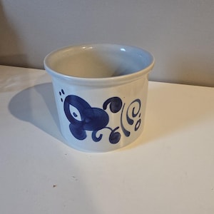 May include: A white ceramic pot with a blue floral design. The cylindrical pot has a slightly flared rim and is decorated with stylized blue flowers and swirls. The pot is sitting on a white surface.